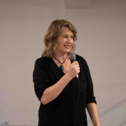 A diretora Tatiana Sager, uma mulher branca de cabelo loiro longo, que veste uma camisa longa de cor preta, fala ao microfone. 

Autoria da descrição da foto: Wagner Meirelles Ascom SJCDH