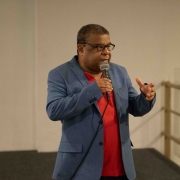 O roteirista Renato Dornelles, um homem negro de óculos, que veste uma camisa social vermelha e um terno azul escuro, fala ao microfone.

Autoria da descrição da foto: Wagner Meirelles Ascom SJCDH