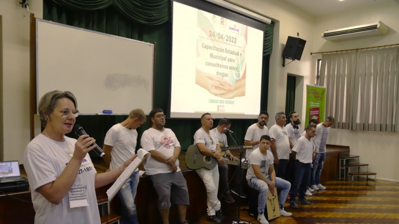 Em frente ao palco do auditório, um grupo de pessoas vestidas de camiseta branca e calças jeans aguarda para iniciar uma apresentação musical que servirá de abertura da capacitação. Dois homens estão segurando violões e um homem está sentado em um cajón - instrumento de madeira que parece uma caixa retangular e que se toca com as mãos. Em cima do palco, há um telão com o pôster do evento em que três pares de mãos estão juntas num gesto de união; acima das mãos está escrito a data do evento "24/04/2023" e o título "Capacitação Estadual e Municipal para conselheiros sobre drogas"; e, embaixo das mãos, está escrito em verde o lema do evento "capacitar para estruturar". No alto do telão, estão as logos dos organizadores: a SJCDH; o Coned-RS e o COMEN-NH.

Autoria da descrição da foto: Wagner Meirelles Ascom SJCDH