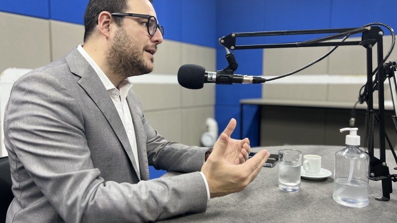 Na imagem, Secretário Mateus Wesp aparece sentado atrás de uma mesa, falando em um microfone. Ele usa óculos, veste um blazer cinza e uma camisa branca. 