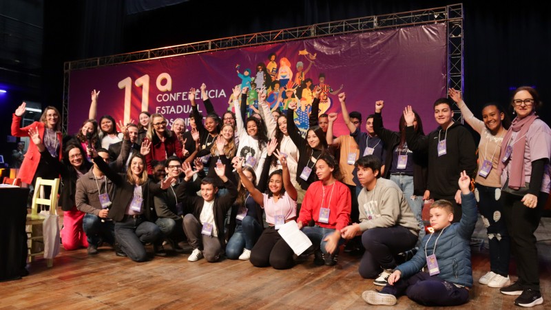Ao fundo da imagem há um banner horizontal roxo escrito 11ª Conferência Estadual dos Direitos da Criança e do Adolescente. Na frente dele, o grupo de delegados do segmento adolescente posa para foto. Há pessoas de pé, com as mãos erguidas e outras agachadas na frente. Eles estão em cima do palco do evento.