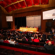 Foto do auditório em que foi realizada a maioria das atividades da Conferência. O público está sentado obervando a mesa de abertura, que está vestida com toalha amarela. 