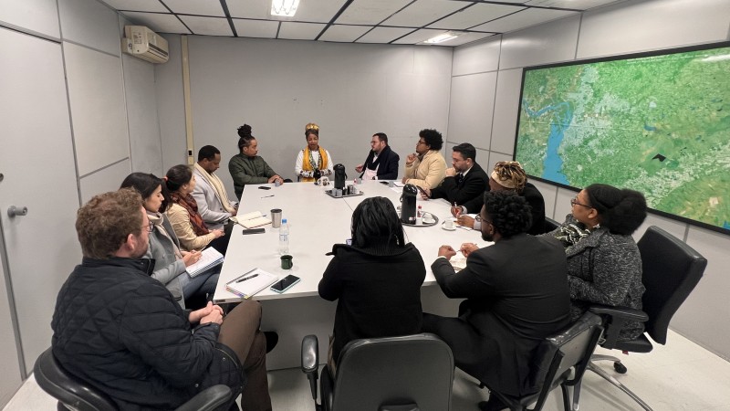 Trezes pessoas estão em volta de uma mesa retangular, na sala de reuniões da SJCDH. A sala não possui janelas e há café e blocos de anotações na mesa. A diretora Sanny está recebendo destaque na foto, ao seu lado direito está o Secretário Mateus Wesp, do esquerdo, o deputado Matheus Gomes. Servidores da Pasta e representantes das deputadas Bruna Rodrigues e Laura Sito também estão presentes.