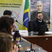 Foto da sala da Comissão de Direitos Humanos na Assembleia Legislativa. Em destaque, o secretário Mateus Wesp, que veste um terno azul escuro. Ele olha para a lateral, em direção a deputada Sofia Cavedon e a Diretora Viviane Viegas. O rosto delas não aparece de frente, a foto foi tirada de perfil. Atrás do Secretário, que está sentado à mesa, há um banner da comissão, de uma instituição e a bandeira do Brasil em um mastro.