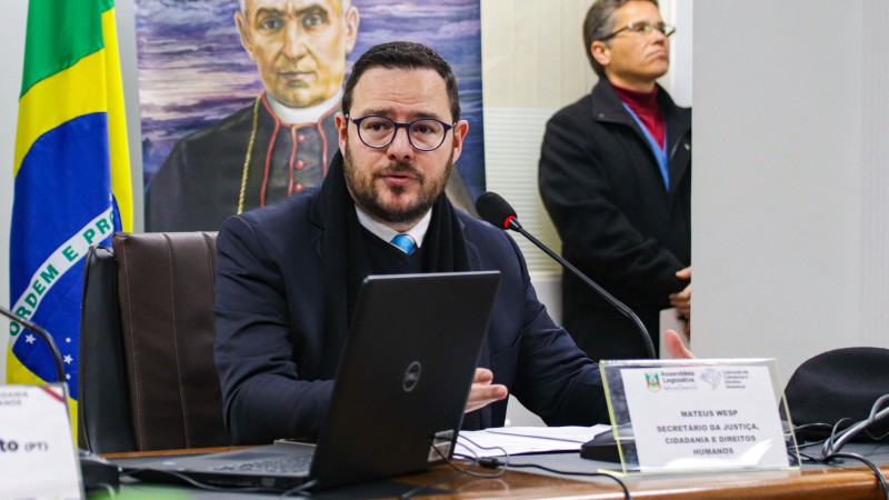 Foto da sala da Comissão de Direitos Humanos na Assembleia Legislativa. Na imagem, aparece o secretário Mateus Wesp, que veste um terno azul escuro. Ele está sentado à mesa, com uma placa transparente com seu nome, um notebook, microfone e papeis. Atrás dele, um banner de uma instituição e a bandeira do Brasil em um mastro. Ao lado do banner há um homem em pé olhando para frente.