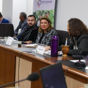Foto da lateral da mesa da sala da comissão. Em destaque, a Diretora Viviane faz uso da palavra. Ela veste um casaco xadrez. Ao seu lado estão o presidente do Comirat, Leonardo Goulart e a deputada Sofia Cavedon, que está com uma térmica e uma cuia de chimarrão a sua frente. 