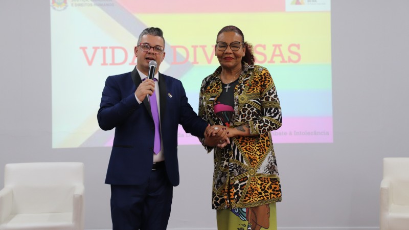 Foto mostra o Diretor do Departamento de Diversidade Sexual, Dani Morethson e a Chefe de Divisão, Glória Crystal no auditório da Fase durante a palestra. Dani e Glória estão de mãos dadas olhando para frente. Dani segura um microfone e veste um terno azul marinho de uma gravata lilás. Glória veste uma saia e um casaco com estampa animal print. Atrás deles há um telão com um slide escrito "Vidas Diversas".