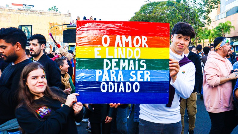 Um casal de jovens com o cartaz com a seguinte frase: “o amor é lindo demais para ser odiado”, com as cores: vermelho, amarelo, verde, azul, e roxo.