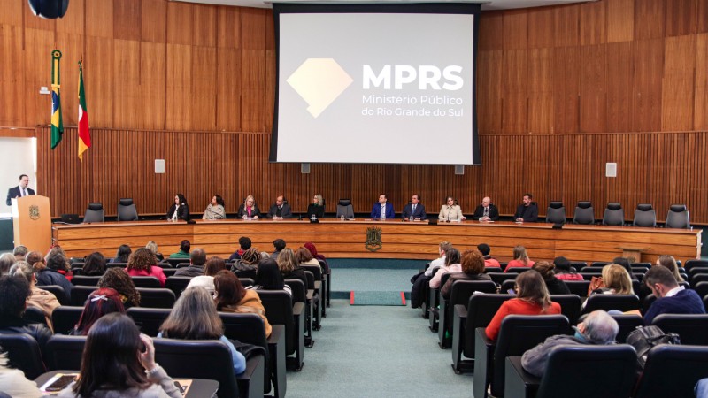 Imagem tirada do fundo do auditório, nela aparece a plateia de costas, sentada. Ao fundo, a mesa de abertura com as autoridades e, acima um telão escrito MPRS Ministério Púbico do Rio Grande do Sul. No púlpito, no canto a esquerda, o secretario Wesp faz uso da palavra.