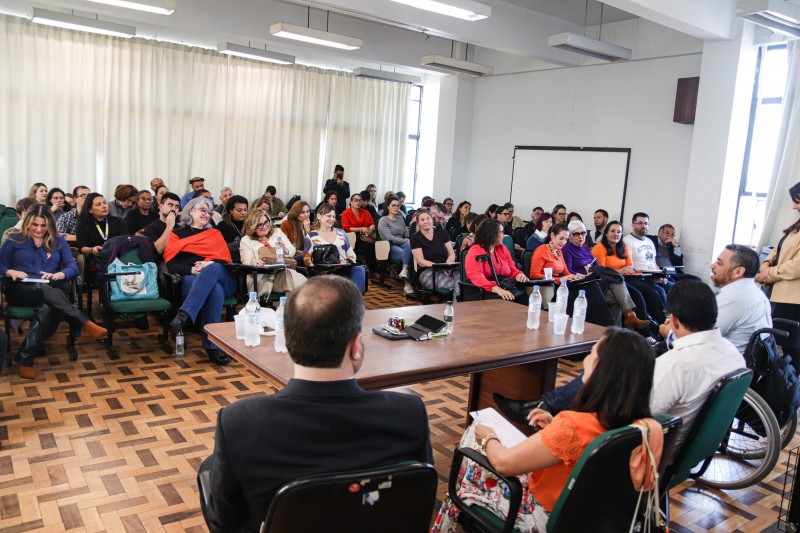 Público observa mesa de abertura durante o Seminário. O público ocupa todas as cadeiras da sala. A sala está pintada com a cor branca com piso de parquê.