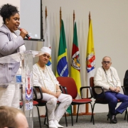Secretaria Adjunta, Caroline Moreira, em seu momento de fala. Ela está em frente ao púlpito. Os demais integrantes do painel observam ela em seu momento de fala.