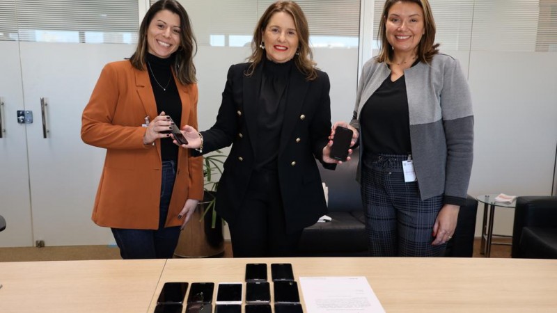 Na foto, a  coordenadora do Centro de Apoio Operacional da Educa&ccedil;&atilde;o, Inf&acirc;ncia e Juventude, Cristiane Della M&eacute;a Corrales posa para foto, ao centro, com a diretora do Departamento de Justi&ccedil;a e Pol&iacute;ticas sobre Drogas, delegada Viviane Viegas, e a analista de Projetos e Pol&iacute;ticas P&uacute;blicas da Divis&atilde;o de Prote&ccedil;&atilde;o &agrave; Vida da SJCDH, Mariele Diotti. Elas est&atilde;o em p&eacute;, atr&aacute;s de uma mesa com 12 celulares. As tr&ecirc;s sorriem para foto enquanto seguram outros dois celulares. 