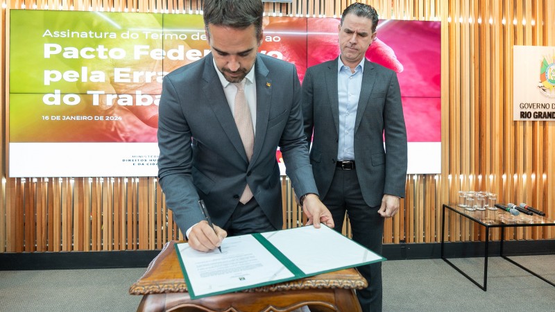 Foto em ambiente fechado. Leite aparece, em plano americano, assinando o documento. Ele está de pé, levemente inclinado sobre uma mesa com o papel. Ao fundo, Peruchin observa o ato.