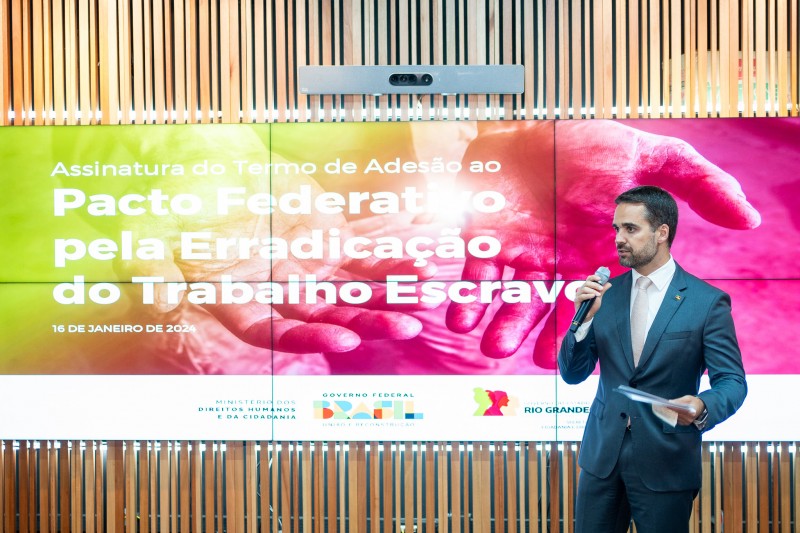 Foto em ambiente fechado, com um fundo de telão colorido com os dizeres "Pacto Federativo pela Erradicação do Trabalho Escravo". Leite aparece à direita, em pé, falando ao microfone que segura na mão.