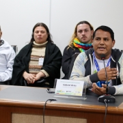 Homem latino, com casaco preto de mangas brancas e azuis. Gesticulando, sentado, em frente ao microfone.