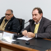 Homem branco, de terno cinza, e blusão amarelo. Sentado falando ao microfone.