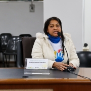 Mulher latina, de casaco branco, camiseta branca, e blusão azul. Sentada falando ao microfone.