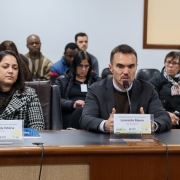Homem branco, de terno cinza e blusão marrom, sentado, gesticulando, falando ao microfone.