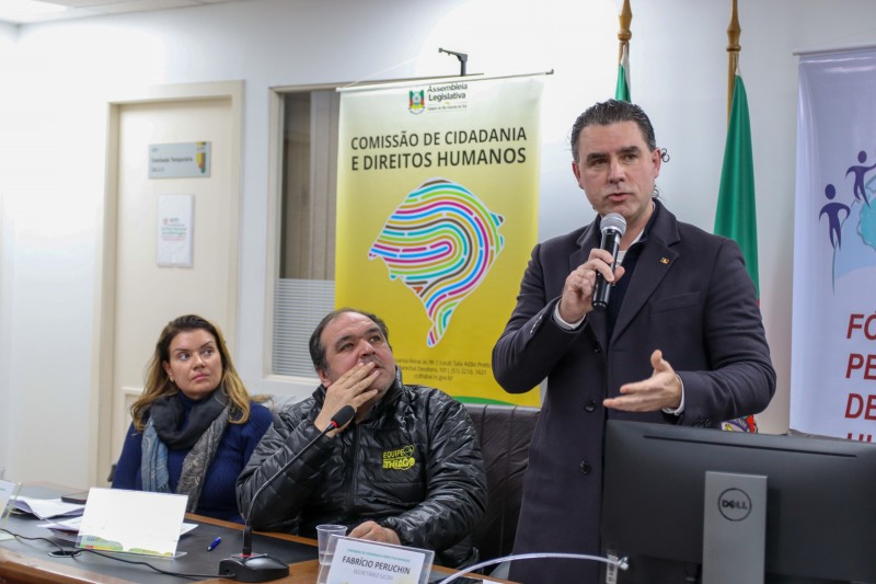 Homem branco, de pé, falando ao microfone, com casaco sobretudo escuro.