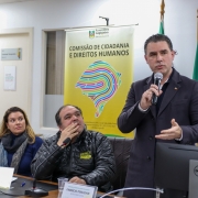 Homem branco, de pé, falando ao microfone, com casaco sobretudo escuro. 
