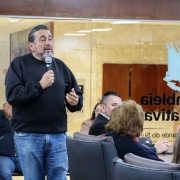 Homem branco, latino, cabelos e barba grisalha, de moletom preto e calça jeans, de pé, com microfone na mão.