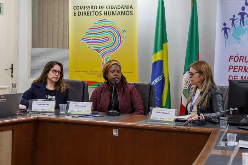 Mulher negra, haitiana, com casaco vermelho, blusa preta, gorro amarelo. Sentada, falando ao microfone.