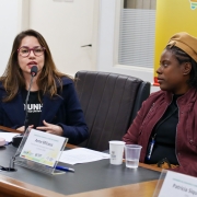 Mulher branca, cabelos castanhos, de óculos, falando ao microfone, sentada, ao lado de Anne Dominique Bruneau, Associação dos Haitianos no Brasil, mulher negra, de casaco vermelho, blusa preta, e gorro amarelo.