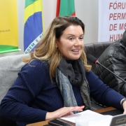 Mulher branca, loira, com casaco azul, e cachecol cinza, sentada, falando ao microfone.