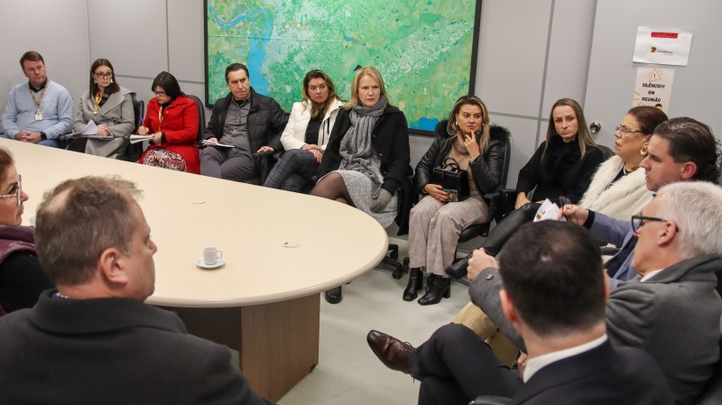 Sala de reuniões com paredes cinzas, com quadro do mapa de Porto Alegre ao fundo e uma mesa elíptica no meio. Homens e mulheres representantes de diferentes poderes públicos sentados lado a lado, ao redor da mesa, com o Secretário da Justiça, Cidadania e Direitos Humanos na ponta.