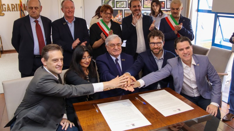 Na imagem, h&aacute; um grupo de onze pessoas reunidas atr&aacute;s de uma das pontas de uma mesa. Algumas est&atilde;o em p&eacute; e outras est&atilde;o sentadas ao redor da mesa de madeira. Sobre a mesa, h&aacute; dois documentos com texto e uma caneta ao lado de cada um, referentes aos pactos firmados pelo munic&iacute;pio de Garibaldi. Cinco pessoas sentadas estendem as m&atilde;os para o centro da mesa, tocando umas nas outras em um gesto simb&oacute;lico de acordo. Ao fundo, h&aacute; quadros pendurados na parede e uma janela parcialmente vis&iacute;vel. Algumas das pessoas em p&eacute; est&atilde;o aplaudindo, indicando a comemora&ccedil;&atilde;o pela assinatura do pacto.