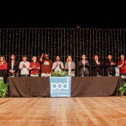 A imagem mostra uma solenidade em um palco amplo, decorado com luzes ao fundo, onde uma longa mesa coberta por uma toalha preta foi posicionada. Sobre ela, há um banner azul com o logotipo do Programa de Oportunidades e Direitos (POD). Em pé atrás da mesa, uma fileira de autoridades e convidados aplaude enquanto olha para o público. Há plantas ornamentais nas extremidades do palco.