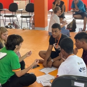 A imagem mostra um grupo de cinco jovens sentados no ch&atilde;o, em c&iacute;rculo, aparentemente participando de uma atividade em grupo ou discuss&atilde;o. Eles est&atilde;o em um espa&ccedil;o interno amplo com piso laranja, que lembra um ambiente de evento, workshop ou din&acirc;mica de grupo. No centro do grupo h&aacute; pap&eacute;is e um celular, sugerindo que est&atilde;o trabalhando em alguma tarefa ou projeto juntos. Um dos jovens, vestindo uma camiseta verde, parece estar explicando algo enquanto gesticula com as m&atilde;os. Os outros quatro est&atilde;o atentos, olhando para ele ou para o centro da roda, indicando que est&atilde;o ouvindo ou debatendo ideias. Alguns deles usam crach&aacute;s pendurados no pesco&ccedil;o, o que refor&ccedil;a a ideia de que participam de um evento organizado, como um curso, treinamento ou encontro estudantil. Ao fundo, h&aacute; outras pessoas tamb&eacute;m sentadas e conversando em pequenos grupos, al&eacute;m de cadeiras e mesas, mostrando que v&aacute;rias atividades semelhantes acontecem no mesmo espa&ccedil;o.