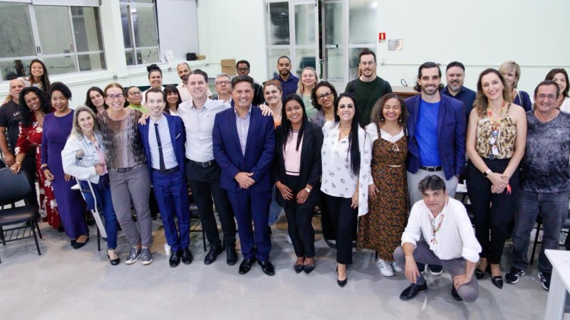 Imagem de um grupo grande de pessoas reunidas em um ambiente interno amplo, audit&oacute;rio universit&aacute;rio da UERGS. As pessoas est&atilde;o posicionadas em p&eacute;, lado a lado, formando uma fileira principal, enquanto outras aparecem atr&aacute;s, compondo um grupo numeroso. Todas est&atilde;o voltadas para a c&acirc;mera. As vestimentas variam entre roupas sociais e casuais, em cores diversas como azul, preto, branco, bege e estampas. Algumas cadeiras pretas aparecem ao fundo e nas laterais do espa&ccedil;o. O ambiente tem paredes claras, piso liso e ilumina&ccedil;&atilde;o intensa. Ao fundo, h&aacute; grandes janelas de vidro, que mostram o exterior escuro, indicando per&iacute;odo noturno. S&atilde;o estudantes, servidores da SJCDH que s&atilde;o professores do curso e autoridades.