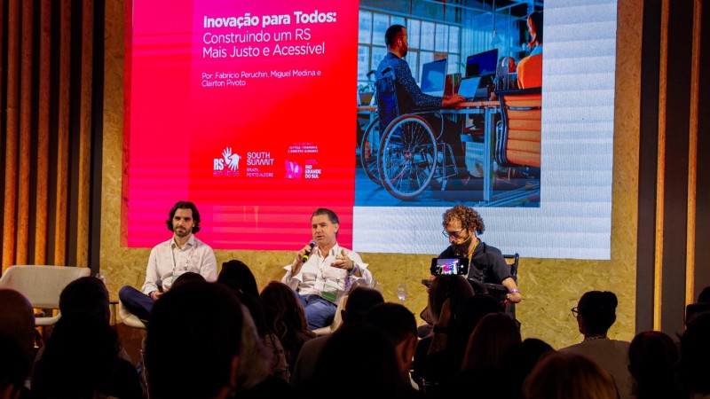 Foto de um painel em um palco, visto a partir da plateia. Tr&ecirc;s pessoas est&atilde;o sentadas em cadeiras sobre o palco; uma delas utiliza cadeira de rodas motorizada. A pessoa ao centro segura um microfone e fala. Ao fundo, um grande tel&atilde;o exibe o t&iacute;tulo &ldquo;Inova&ccedil;&atilde;o para Todos: Construindo um RS Mais Justo e Acess&iacute;vel&rdquo;, com imagem de uma pessoa em cadeira de rodas em ambiente de trabalho. A plateia aparece em primeiro plano, sentada e voltada ao palco. O cen&aacute;rio tem ilumina&ccedil;&atilde;o profissional e acabamento em madeira.
