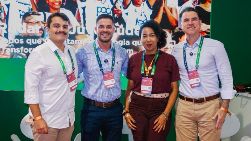 Quatro pessoas, uma mulher negra e tr&ecirc;s homens brancos, est&atilde;o posicionadas lado a lado em um espa&ccedil;o de evento, usando crach&aacute;s pendurados no pesco&ccedil;o e roupas sociais, como camisas e cal&ccedil;as. Ao fundo, h&aacute; um painel colorido com imagens de p&uacute;blico e texto parcialmente vis&iacute;vel, incluindo &ldquo;Radar da Juventude&rdquo;. &Eacute; o estande do governo no evento, com ilumina&ccedil;&atilde;o uniforme e estrutura de apresenta&ccedil;&atilde;o. As pessoas est&atilde;o centralizadas na imagem, ocupando o primeiro plano diante do painel institucional.