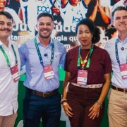 Quatro pessoas, uma mulher negra e tr&ecirc;s homens brancos, est&atilde;o posicionadas lado a lado em um espa&ccedil;o de evento, usando crach&aacute;s pendurados no pesco&ccedil;o e roupas sociais, como camisas e cal&ccedil;as. Ao fundo, h&aacute; um painel colorido com imagens de p&uacute;blico e texto parcialmente vis&iacute;vel, incluindo &ldquo;Radar da Juventude&rdquo;. &Eacute; o estande do governo no evento, com ilumina&ccedil;&atilde;o uniforme e estrutura de apresenta&ccedil;&atilde;o. As pessoas est&atilde;o centralizadas na imagem, ocupando o primeiro plano diante do painel institucional.