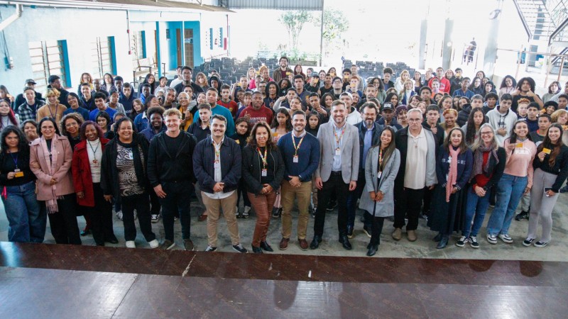 Grupo numeroso de pessoas posa em um espa&ccedil;o amplo coberto, semelhante a um p&aacute;tio escolar ou audit&oacute;rio aberto. Na frente, uma fileira de adultos com crach&aacute;s, servidores da SJCDH; atr&aacute;s, muitos jovens ocupam cadeiras e ficam em p&eacute;. Algumas pessoas seguram cart&otilde;es amarelos. Ao fundo h&aacute; colunas, escadas met&aacute;licas e &aacute;reas com vegeta&ccedil;&atilde;o, com luz natural intensa entrando pelo lado direito.
