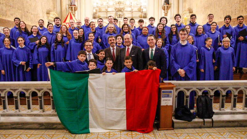 Grupo numeroso posa dentro de uma igreja decorada, com altar ao fundo e detalhes dourados. A maioria usa t&uacute;nicas azuis e segura pastas. Na frente, pessoas exibem uma grande bandeira da It&aacute;lia, nas cores verde, branco e vermelho. Alguns trajes sociais aparecem entre o grupo. O ambiente &eacute; iluminado e possui grades de pedra na parte frontal.