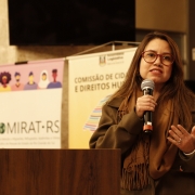 Mulher branca de cabelos escuros posicionada atrás de um púlpito de madeira segura um microfone com a mão direita e gesticula com a outra. Usa óculos e veste casaco e cachecol em tom neutro. Ao fundo, banners exibem “Comissão de Cidadania e Direitos Humanos” e “Comirat-RS”, além de ilustrações de pessoas. Bandeira aparece à direita. Ambiente interno, com iluminação quente.