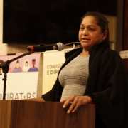 Mulher de cabelos escuros e presos, em pé atrás de um púlpito de madeira fala ao microfone acoplado a um suporte. Veste peça escura sobre roupa clara e mantém uma das mãos apoiada no púlpito. Ao fundo, banners exibem “Comissão de Cidadania e Direitos Humanos” e “Comirat-RS”, com ilustrações de pessoas. À direita, uma bandeira vertical com laço verde, amarelo e vermelho. Ambiente interno com iluminação quente e parede de madeira.