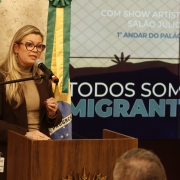 Mulher branca de cabelos claros posicionada atrás de um púlpito de madeira fala ao microfone preso a um suporte. Usa óculos, veste blazer escuro sobre blusa clara e mantém uma das mãos levemente levantada, em gesto. Ao fundo, duas faixas verde e amarela e um telão com o texto “TODOS SOMOS MIGRANTES”. À esquerda, parte de uma bandeira. Ambiente interno com iluminação quente.