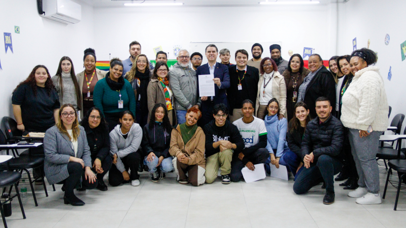 Foto de grupo feita em uma sala de reuni&otilde;es iluminada por luz branca. Cerca de 30 pessoas est&atilde;o reunidas para um registro coletivo, distribu&iacute;das em tr&ecirc;s fileiras: algumas em p&eacute; ao fundo, outras agachadas ou ajoelhadas &agrave; frente. No centro, duas pessoas seguram um documento voltado para a c&acirc;mera. O ambiente possui paredes brancas, cadeiras escolares nas laterais e cartazes coloridos ao fundo, incluindo faixas com as cores do arco-&iacute;ris. H&aacute; participantes usando crach&aacute;s e roupas de inverno, como casacos, cachec&oacute;is e blus&otilde;es. A imagem transmite um momento de encontro institucional e participa&ccedil;&atilde;o coletiva.