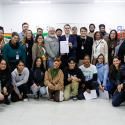 Foto de grupo feita em uma sala de reuni&otilde;es iluminada por luz branca. Cerca de 30 pessoas est&atilde;o reunidas para um registro coletivo, distribu&iacute;das em tr&ecirc;s fileiras: algumas em p&eacute; ao fundo, outras agachadas ou ajoelhadas &agrave; frente. No centro, duas pessoas seguram um documento voltado para a c&acirc;mera. O ambiente possui paredes brancas, cadeiras escolares nas laterais e cartazes coloridos ao fundo, incluindo faixas com as cores do arco-&iacute;ris. H&aacute; participantes usando crach&aacute;s e roupas de inverno, como casacos, cachec&oacute;is e blus&otilde;es. A imagem transmite um momento de encontro institucional e participa&ccedil;&atilde;o coletiva.